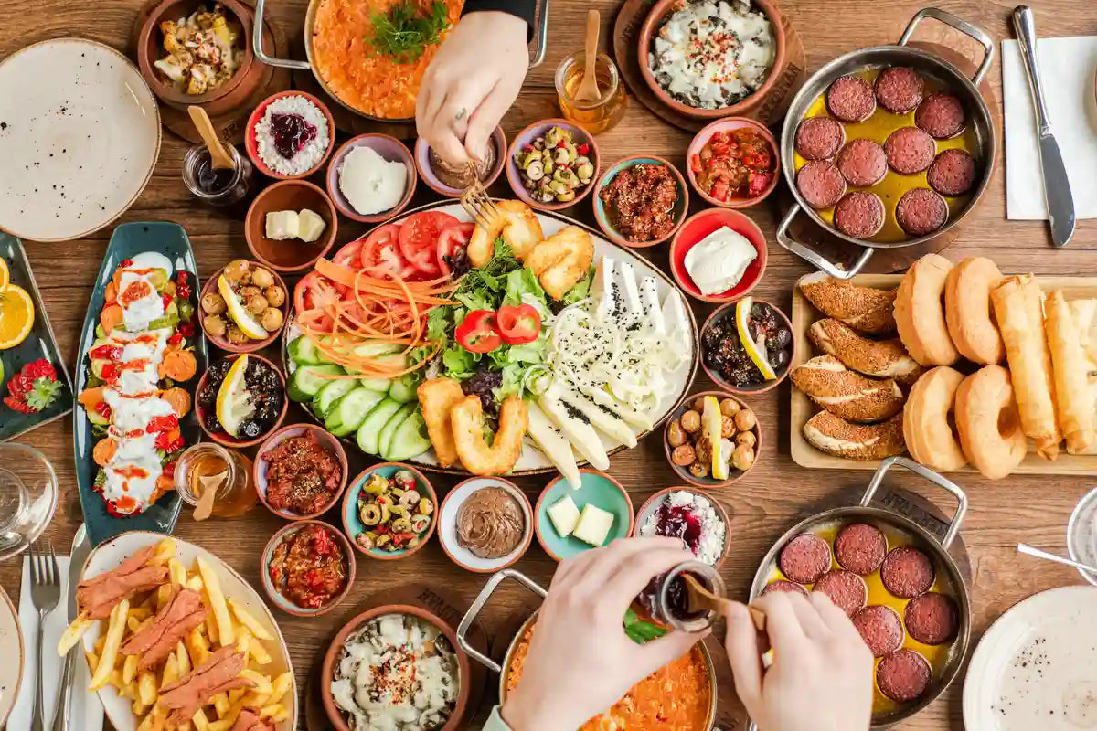 table full of food like salad, fries, pastries, and more, and two people's hands are seen taking food