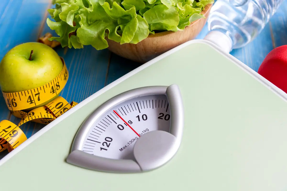 weighing scale next to an apple wrapped with a tape measure, lettuce in a bowl, and bottled water